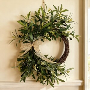 May include: An olive branch wreath with a natural brown twig base. The wreath features green leaves, black olives, and small white flowers. A beige burlap bow adds a rustic touch. Ideal for home decoration.