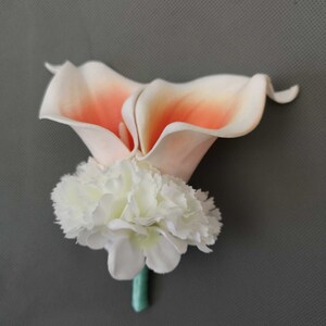 May include: A peach and white artificial flower boutonniere with a single white carnation and two peach calla lilies.