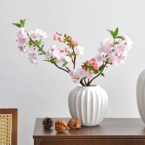 May include: A white, ribbed ceramic vase holds branches of artificial cherry blossoms. The blossoms are a mix of pink and white, with green leaves. The vase sits on a dark brown surface with decorative pinecones and nuts.