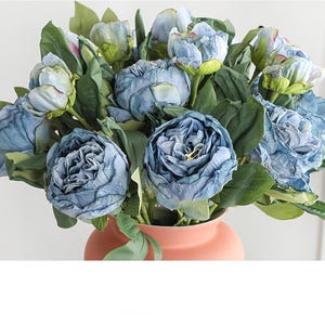 May include: A bouquet of blue artificial peonies with green leaves in a peach-colored vase. The flowers have a ruffled texture and are in various stages of bloom, creating a full and lush arrangement. The vase is a simple, rounded shape.