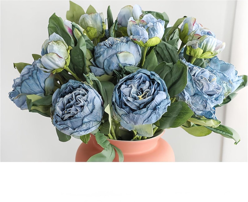 May include: A bouquet of blue artificial peonies with green leaves in a peach-colored vase. The flowers have a ruffled texture and are in various stages of bloom, creating a full and lush arrangement. The vase is a simple, rounded shape.
