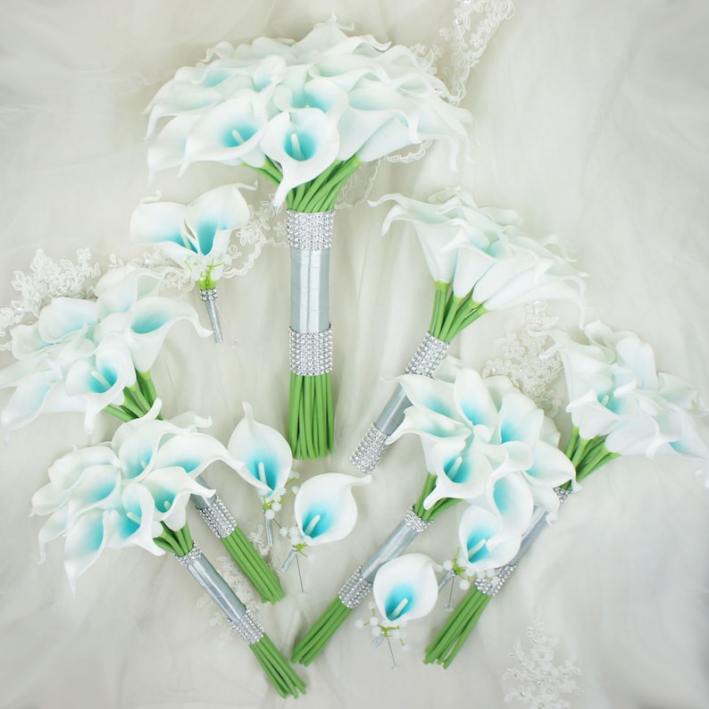 May include: A set of six white calla lily bouquets with blue accents. The bouquets are tied with silver ribbon and rhinestones. The bouquets are arranged on a white fabric background.