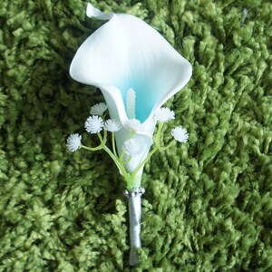 May include: A white calla lily with a light blue center, accented with small white flowers, is attached to a silver stem. The bouquet is perfect for a wedding or special occasion.