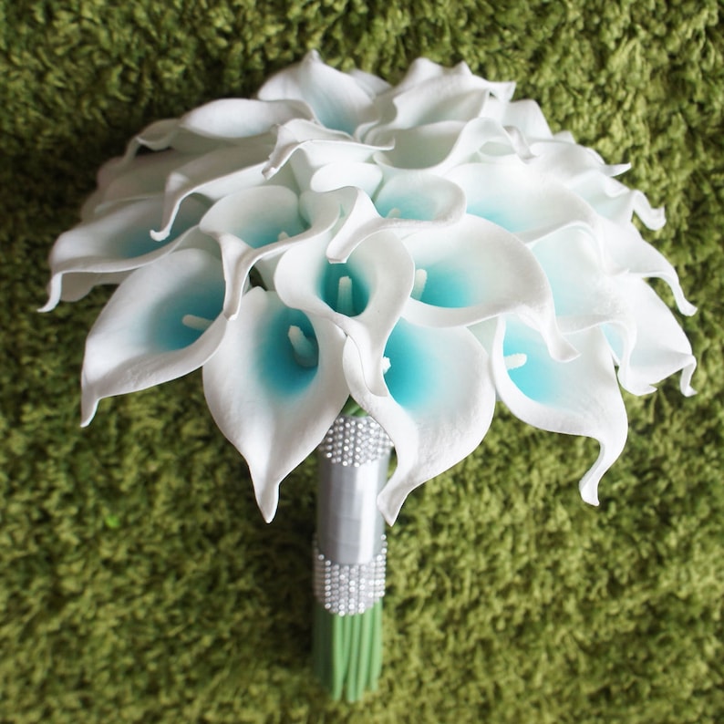 May include: A white bridal bouquet made of artificial calla lilies with blue accents. The bouquet is tied with a silver ribbon and rhinestones.