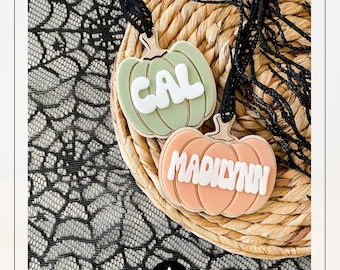 May include: Two pumpkin-shaped ornaments with names, "CAL" and "MADILYNN", in white block letters. The pumpkins are light green and peach, with a wooden stem. The background includes a woven basket and black lace.