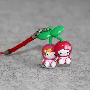May include: A cherry-themed charm featuring two Hello Kitty figures. The figures are dressed in pink and red, with a green stem and leaves. The charm has a red braided cord and a silver clasp.
