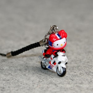 May include: A small, colorful Hello Kitty figurine riding a white motorcycle. The figurine is wearing a red racing suit and helmet. The motorcycle has blue and red accents. A black lanyard is attached.