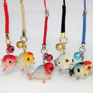 Op de afbeelding: Vijf telefoonhangers met miniatuur goudvisfiguurtjes in diverse kleuren zoals rood, wit en blauw, elk bevestigd aan een gevlochten koord met een klein metalen belletje. De goudvissen hebben gedetailleerde ogen en vinnen en hangen aan goud- of zilverkleurige metalen ringen.