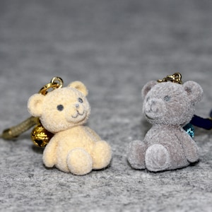 Könnte beinhalten: Zwei kleine Plüsch-Teddybär-Schlüsselanhänger. Einer ist beige und der andere grau. Beide haben goldfarbene Ringe und sind an Schnüren befestigt.