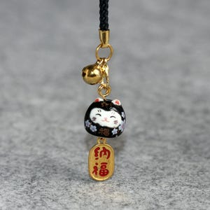 Puede incluir: Un amuleto de gato de cerámica negro y blanco con una campana dorada y un amuleto dorado con caracteres japoneses que se leen "招福" (que significa "invitar la buena fortuna").