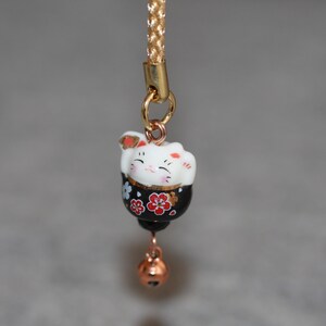 May include: A black and white ceramic lucky cat charm with red and gold accents. The charm is attached to a gold chain with a gold bell.