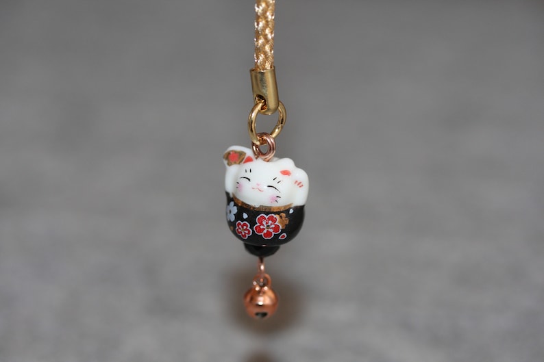 May include: A black and white ceramic lucky cat charm with red and gold accents. The charm is attached to a gold chain with a gold bell.