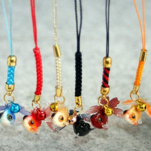 Peut inclure: Six breloques pour téléphone avec des figurines de poissons rouges miniatures en bleu, rouge, blanc, noir, rouge foncé et orange. Chaque breloque possède un cordon tressé, un connecteur en métal doré et une petite clochette sur le poisson.