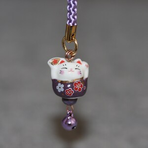 May include: A purple and white ceramic Japanese Maneki-Neko, or lucky cat, charm with a bell. The cat is holding its right paw up and has a gold design on its chest. The charm is attached to a purple cord.