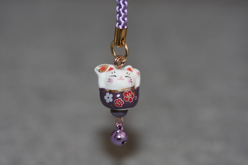 May include: A purple and white ceramic Japanese Maneki-Neko, or lucky cat, charm with a bell. The cat is holding its right paw up and has a gold design on its chest. The charm is attached to a purple cord.