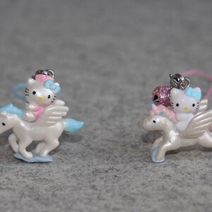 May include: Two Hello Kitty charms featuring Hello Kitty riding a white winged unicorn. The unicorn has a blue tail and mane. The charms are pink and blue.