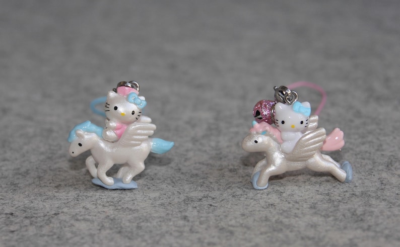 May include: Two Hello Kitty charms featuring Hello Kitty riding a white winged unicorn. The unicorn has a blue tail and mane. The charms are pink and blue.