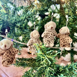 Könnte beinhalten: Vier kleine Teddybär-Ornamente hängen an einem Weihnachtsbaum. Die Bären tragen braune und beige gemusterte Outfits. Jedes Ornament hat einen silberfarbenen Verschluss und eine schwarze Schnur zum Aufhängen.