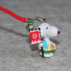 Pode incluir: Uma figurinha branca do Snoopy vestindo uma roupa azul e verde, segurando um objeto triangular marrom. O Snoopy está carregando uma bandeira vermelha e rosa com caracteres japoneses. A figurinha está presa a um cordão vermelho com um sino verde.