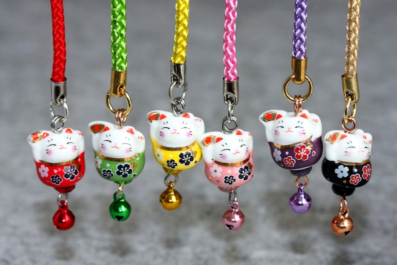 May include: Seven lucky cat charms with different colored bodies and bells. The charms are attached to cords of different colors. The charms are all in a row and are facing the camera.