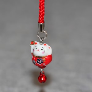 May include: A red and white ceramic lucky cat charm with black floral details, hanging from a red string with a silver bell.