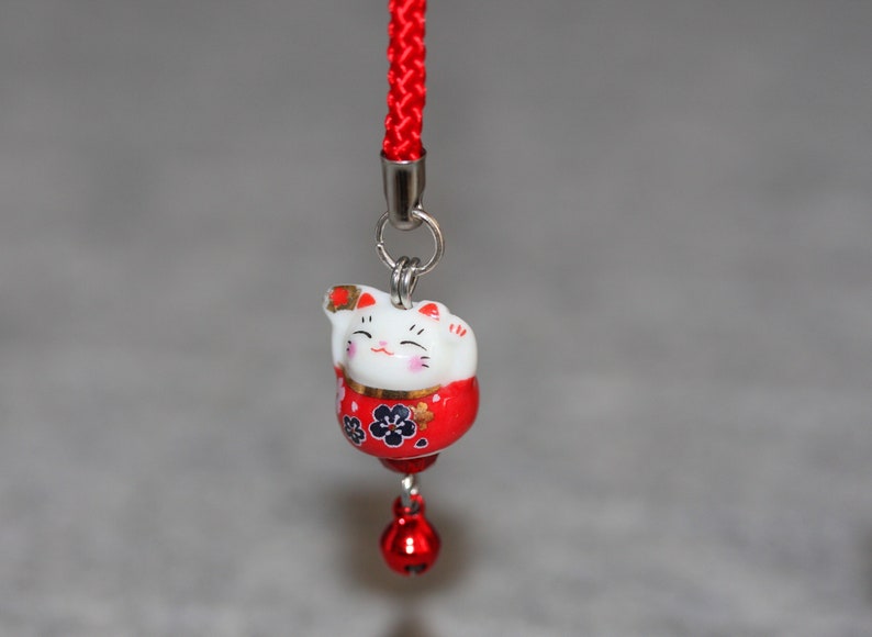 May include: A red and white ceramic lucky cat charm with black floral details, hanging from a red string with a silver bell.