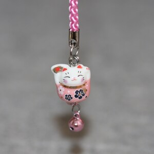 May include: A pink and white ceramic Maneki-Neko, or lucky cat, charm with black and gold floral details. The charm is attached to a pink and white cord with a silver bell.