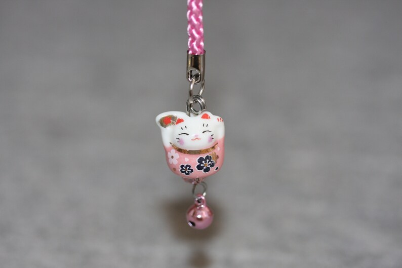 May include: A pink and white ceramic Maneki-Neko, or lucky cat, charm with black and gold floral details. The charm is attached to a pink and white cord with a silver bell.