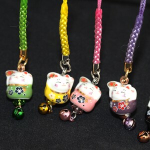 May include: Seven colorful Japanese Maneki-Neko, or lucky cat, charms with bells. Each charm is a different color: red, green, yellow, pink, purple, black, and white. The charms are attached to cords of matching colors.