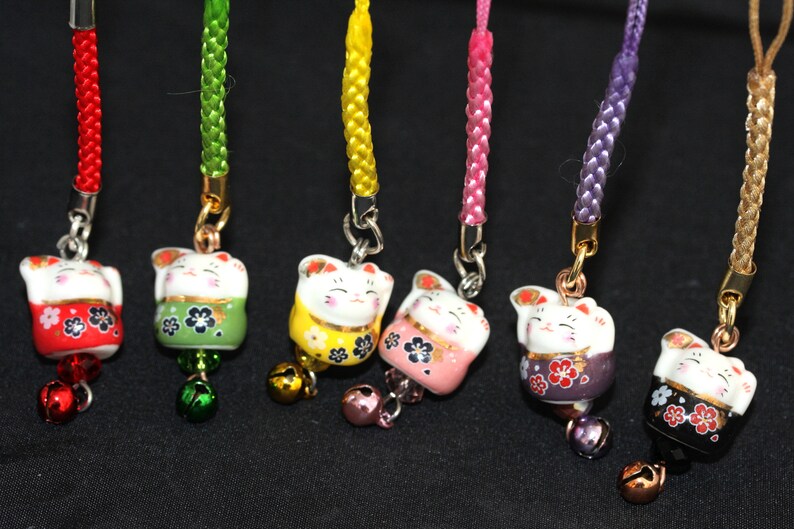 May include: Seven colorful Japanese Maneki-Neko, or lucky cat, charms with bells. Each charm is a different color: red, green, yellow, pink, purple, black, and white. The charms are attached to cords of matching colors.