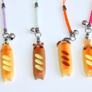 May include: Four novelty phone charms shaped like animals on bread. The charms include a cat, bear, dog, and bunny, each with a bell and colourful cord. The bread is orange and brown, with yellow accents.