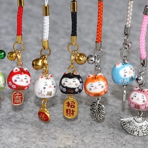 Puede incluir: Siete amuletos japoneses Maneki-Neko, o gatos de la suerte, de colores y diseños diferentes. Cada amuleto está unido a una cuerda con una campanilla y un elemento decorativo. Los amuletos están colgados de una superficie blanca.