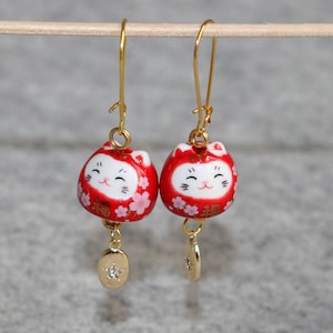 May include: Gold earrings with red and white ceramic charms featuring a smiling cat face and the Japanese word "福" (meaning "good fortune").