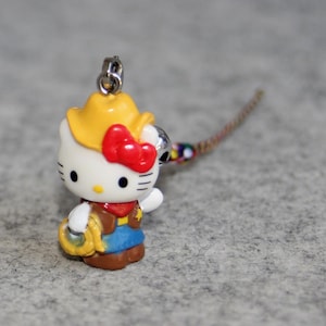 May include: A white Hello Kitty figurine wearing a yellow cowboy hat and a red bandana. She is holding a lasso in her right hand and is wearing a blue and brown vest. The figurine is attached to a silver chain.