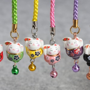May include: Seven colorful Japanese Maneki-Neko, or lucky cat, charms with bells. Each charm features a white cat with a different colored body and floral pattern. The charms are attached to cords of matching colors.