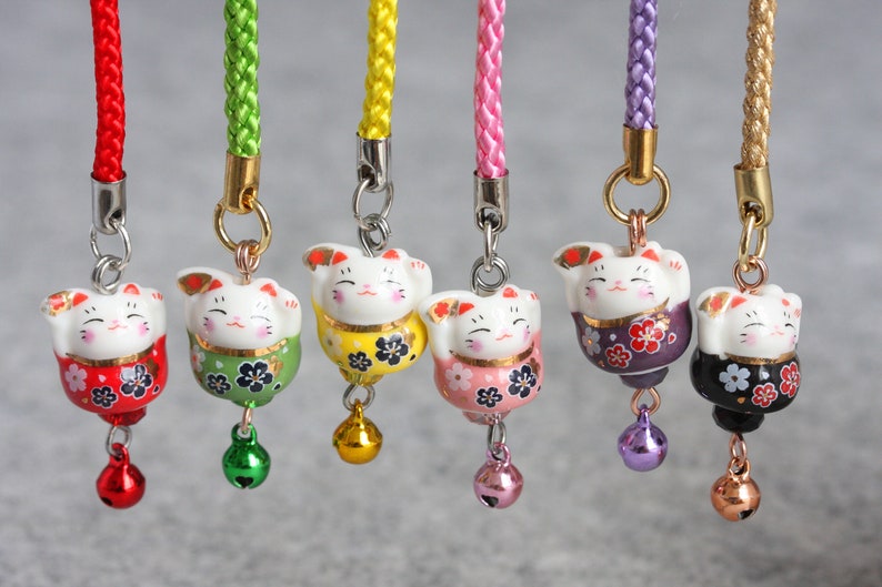 May include: Seven colorful Japanese Maneki-Neko, or lucky cat, charms with bells. Each charm features a white cat with a different colored body and floral pattern. The charms are attached to cords of matching colors.