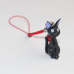 Può includere: Un ciondolo a forma di gatto nero con orecchie blu e grandi occhi bianchi, attaccato a un cordino rosso. Il gatto è seduto in posizione eretta, con una piccola campana sulla testa. Un accessorio carino.