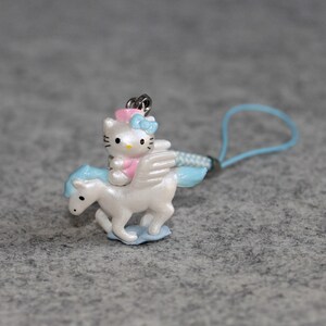 May include: A white and blue winged unicorn charm with a pink and blue Hello Kitty riding on its back. The charm is attached to a light blue string.