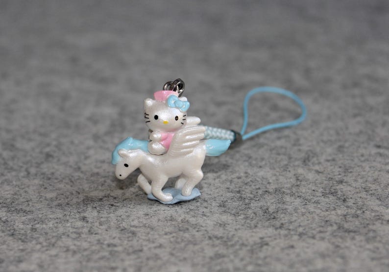 May include: A white and blue winged unicorn charm with a pink and blue Hello Kitty riding on its back. The charm is attached to a light blue string.