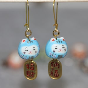 Pode incluir: Um par de brincos dourados com amuletos de gato da sorte em cerâmica azul clara. Cada gato tem um rosto sorridente, detalhes florais e um pingente de moeda dourada com caracteres vermelhos. Os brincos penduram-se em fios dourados.