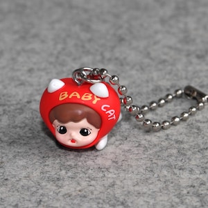 Könnte beinhalten: Ein roter und weißer Schlüsselanhänger mit einem Cartoon-Baby in einem Katzenkostüm. Der Anhänger trägt die Aufschrift "BABY CAT". Er ist an einer silbernen Kugelkette befestigt.