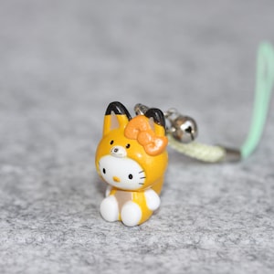 May include: A small figurine charm featuring a white cat character wearing a yellow fox hood with black-tipped ears and a matching yellow bow. The charm includes a small silver-colored metal bell and a light green cord loop for attachment.