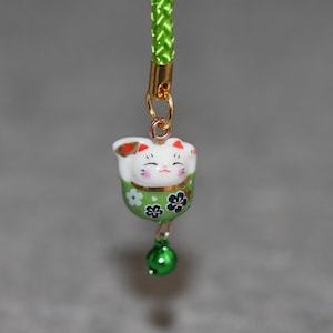 May include: A green and white ceramic lucky cat charm with a gold bell hanging from it. The charm is attached to a green cord.
