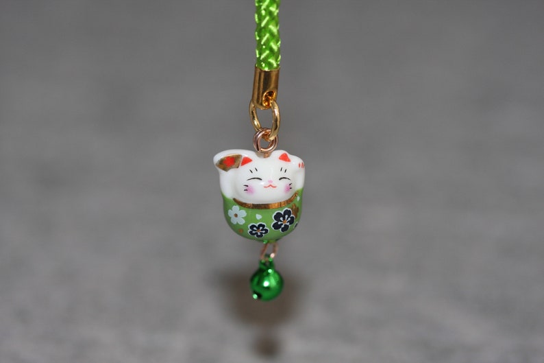 May include: A green and white ceramic lucky cat charm with a gold bell hanging from it. The charm is attached to a green cord.
