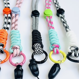 Op de afbeelding: Vijf kleurrijke lanyard-sleutelhangers met zwarte sluitingen. De lanyards zijn gemaakt van gevlochten touw en hebben verschillende kleurcombinaties, waaronder zwart-wit, oranje-wit, roze-blauw, zwart-wit en roze-groen.