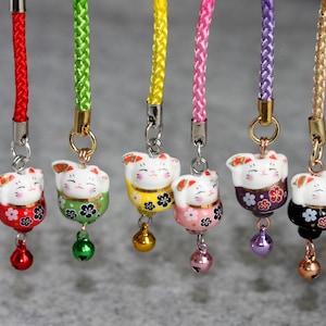 May include: Seven lucky cat charms with different colored cords and bells. The cats are white with painted details and are holding up their right paw. The charms are on a white background.