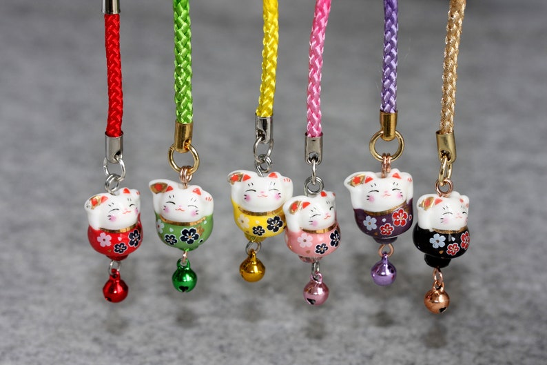 May include: Seven lucky cat charms with different colored cords and bells. The cats are white with painted details and are holding up their right paw. The charms are on a white background.
