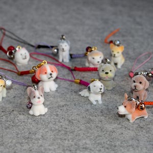Puede incluir: Una colección de pequeñas figuras de perros de colores con cordones adjuntos. Los perros son de varias razas y colores, incluyendo blanco, beige, gris y marrón. Cada figura tiene una pequeña campana y un cordón decorativo.
