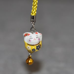 May include: A yellow and white ceramic charm of a smiling cat with black flowers on its chest. The charm is attached to a yellow string with a silver bell.