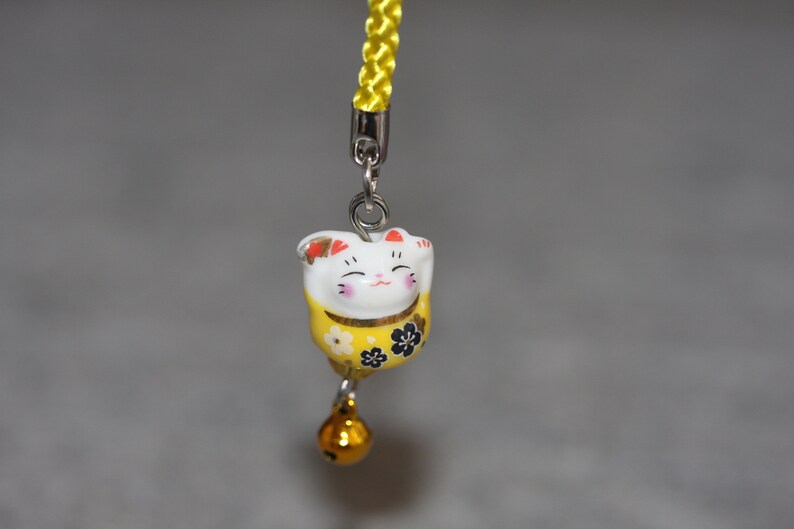 May include: A yellow and white ceramic charm of a smiling cat with black flowers on its chest. The charm is attached to a yellow string with a silver bell.